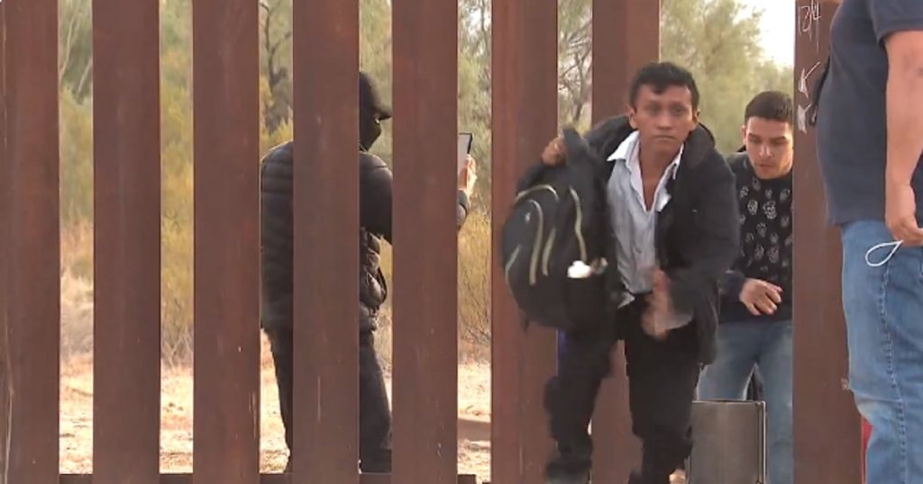 Illegal aliens pour through a breach in the border wall in Lukeville, Arizona, while a human smuggler records their faces.